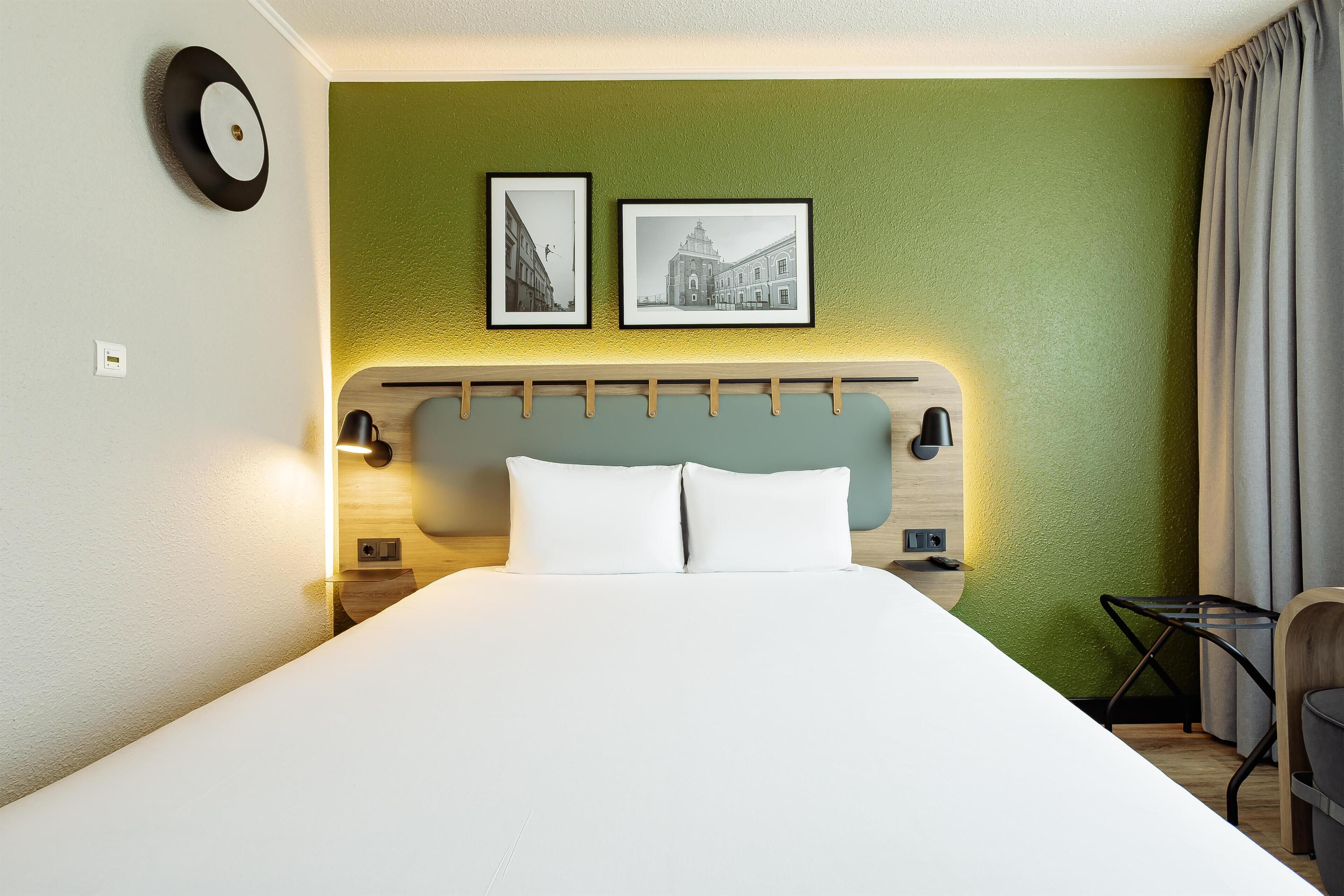 A hotel room with a bed, a headboard, two framed pictures, a nightstand, a lamp, a chair, and a curtain.