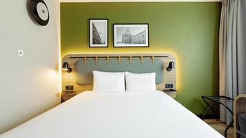 A hotel room with a bed, a headboard, two framed pictures, a nightstand, a lamp, a chair, and a curtain.