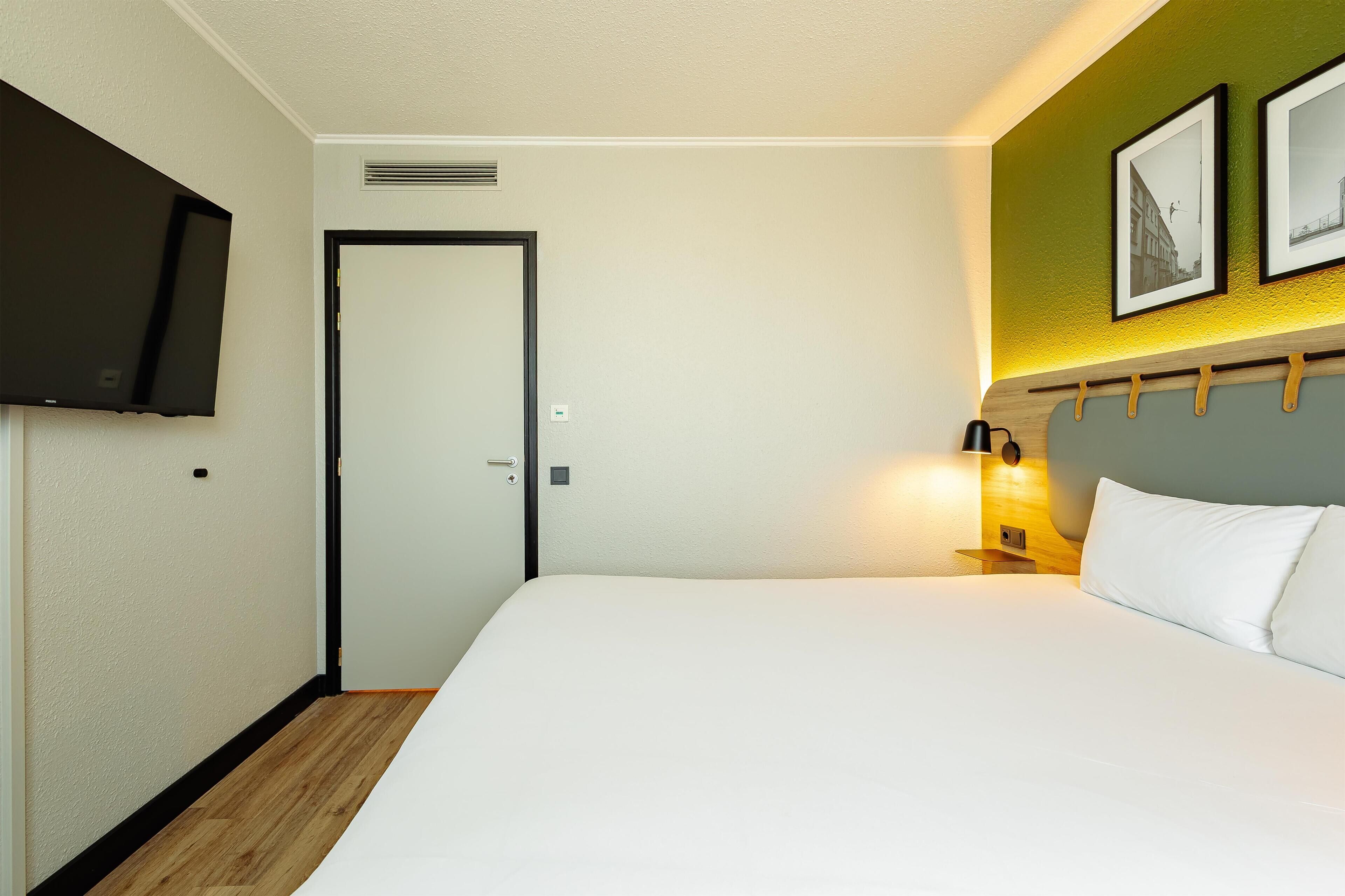 A hotel room with a bed, a television mounted on the wall, a door, and a wall-mounted light fixture.
