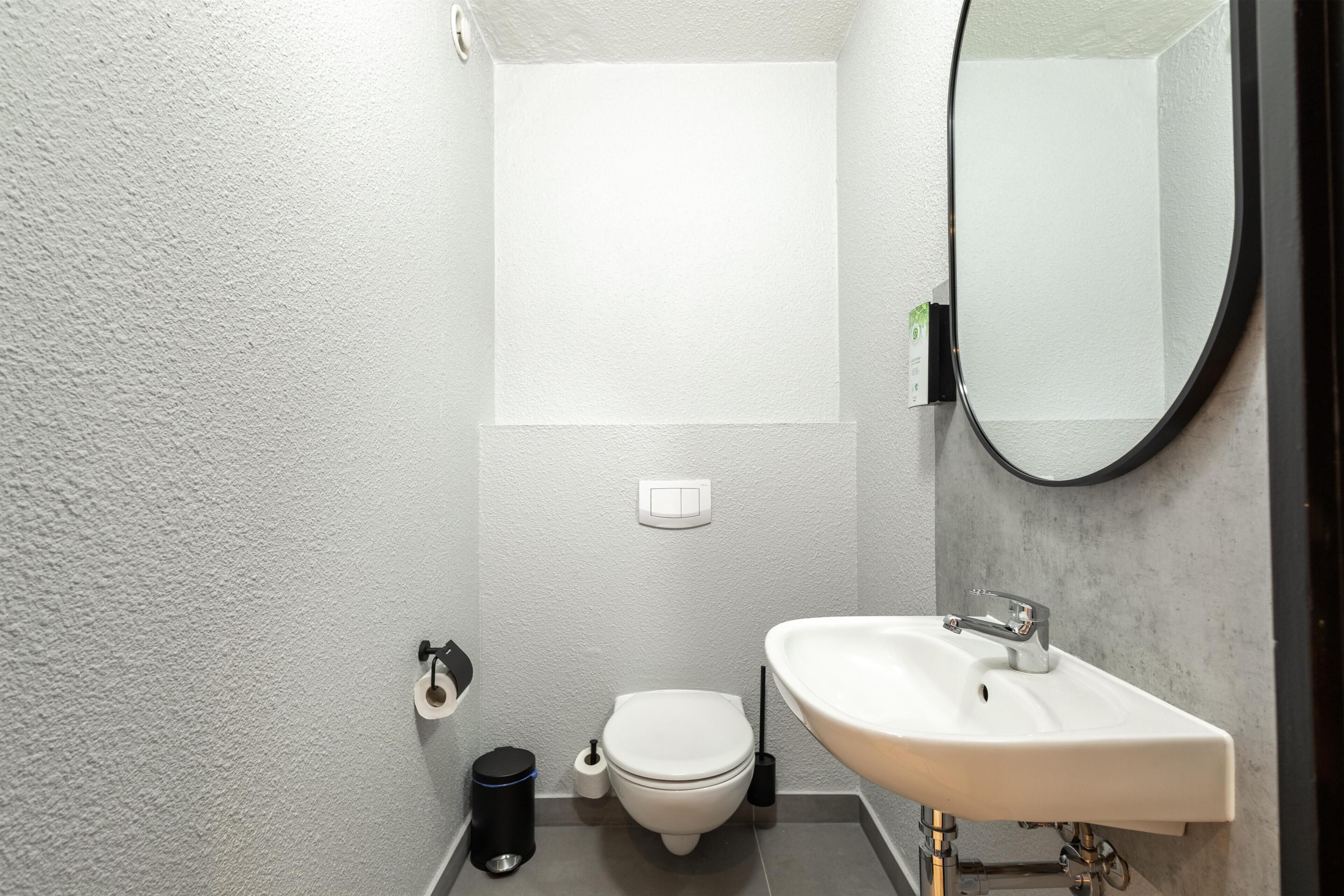A bathroom with a toilet, sink, mirror, and a trash bin.
