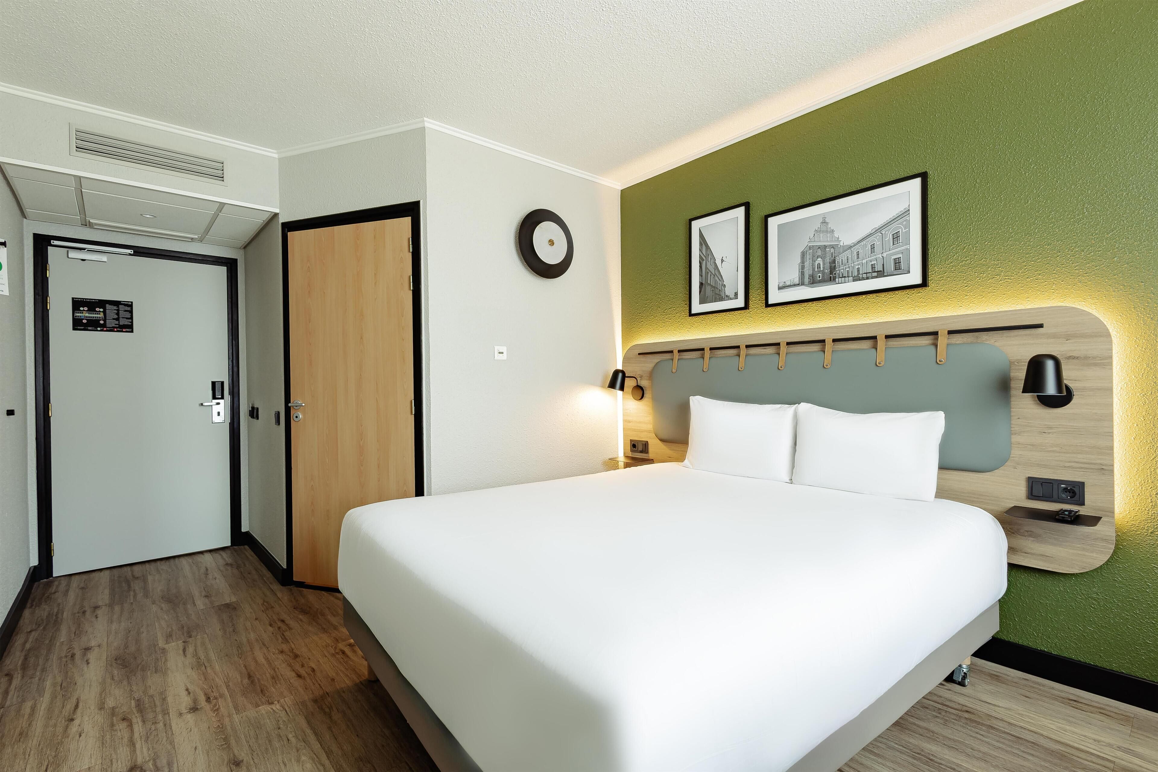A hotel room with a bed, a wooden headboard, a wall-mounted clock, and two framed pictures.