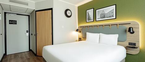 A hotel room with a bed, a wooden headboard, a wall-mounted clock, and two framed pictures.