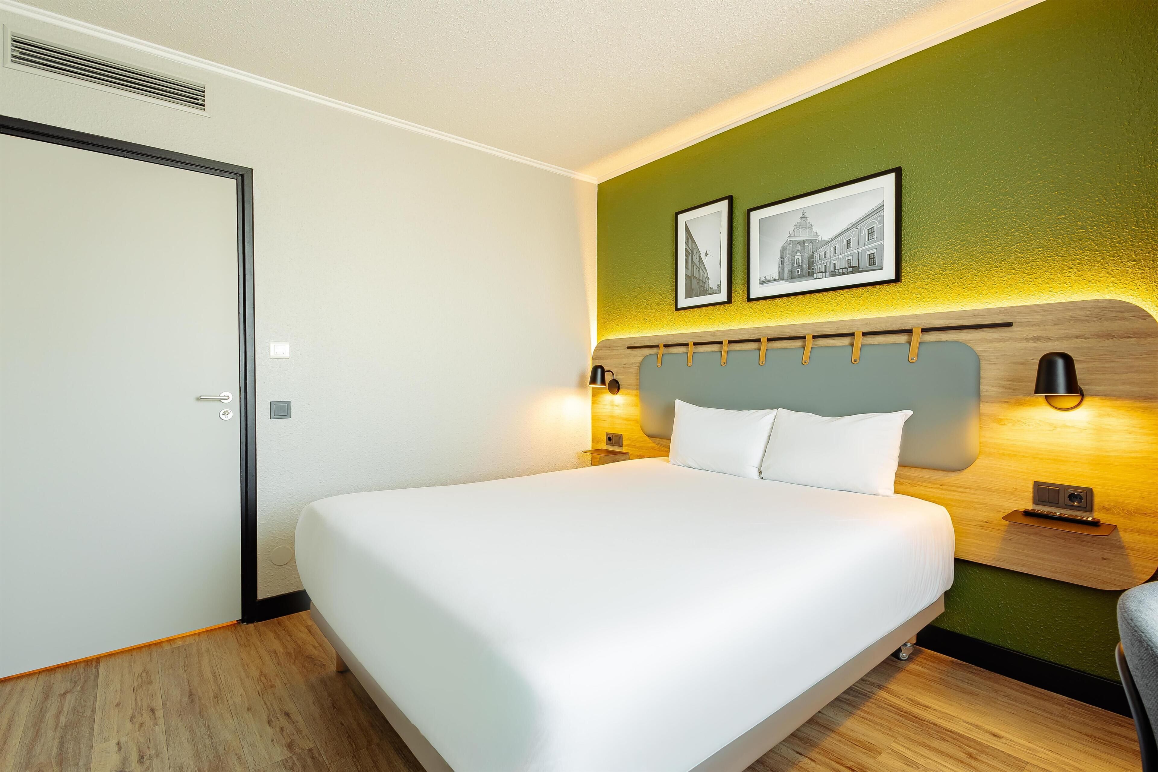 Una habitación de hotel con una cama grande, un cabecero de madera, dos lámparas fijadas a la pared y una puerta.