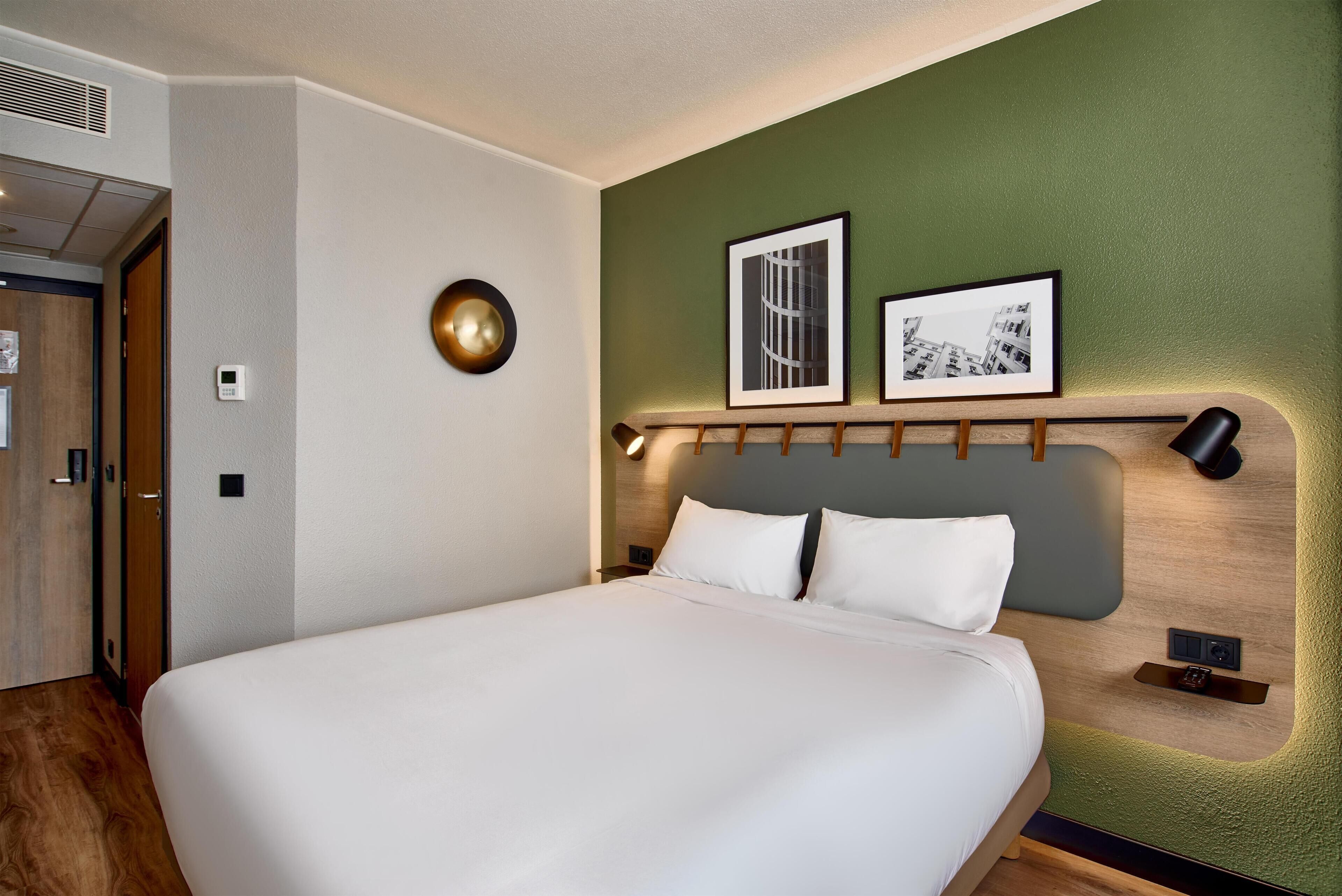 Una habitación de hotel moderna con una cama grande, un cabecero de madera, una pared de acento verde y cuadros enmarcados.