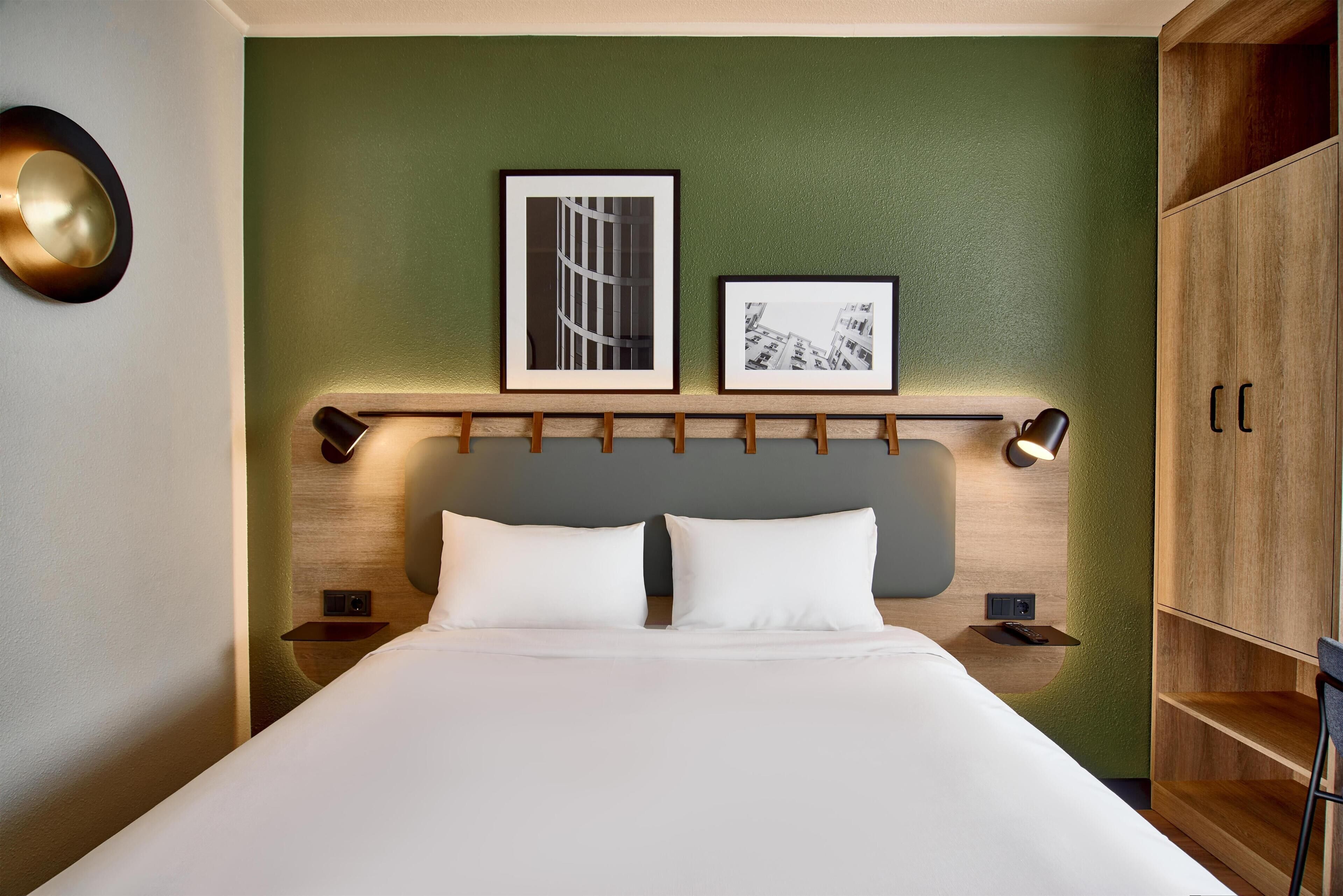 A neatly made bed with white linens, a green accent wall, and two framed pictures above it.