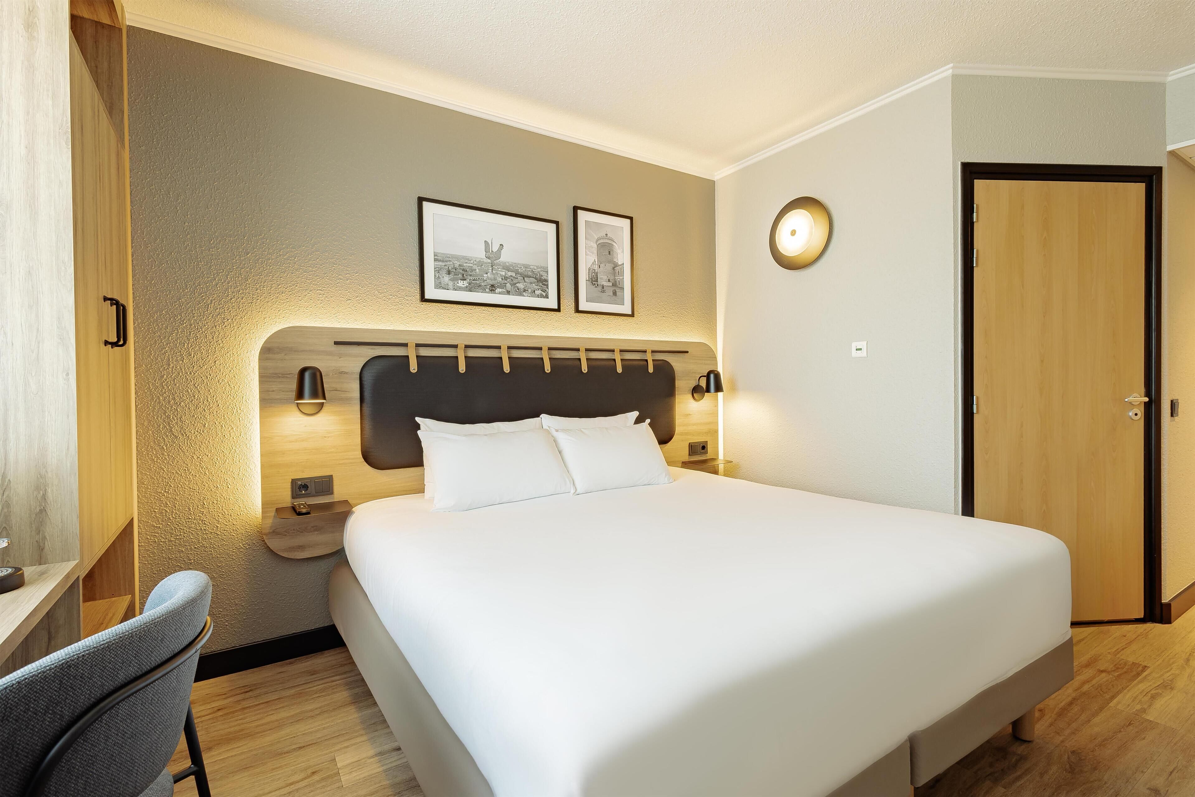 Una habitación de hotel con una cama grande, un escritorio con silla, una mesita de noche, una lámpara de pared y una puerta.