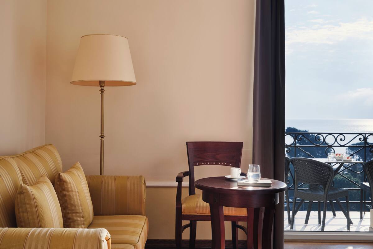 A hotel room with a sofa, a chair, a table with a cup and glass, and a view of the sea through a balcony.