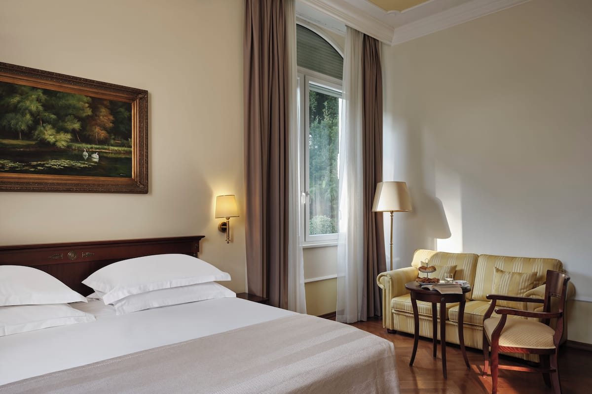 A hotel room with a large bed, a painting, a sofa, a chair, a lamp, and a window with curtains.