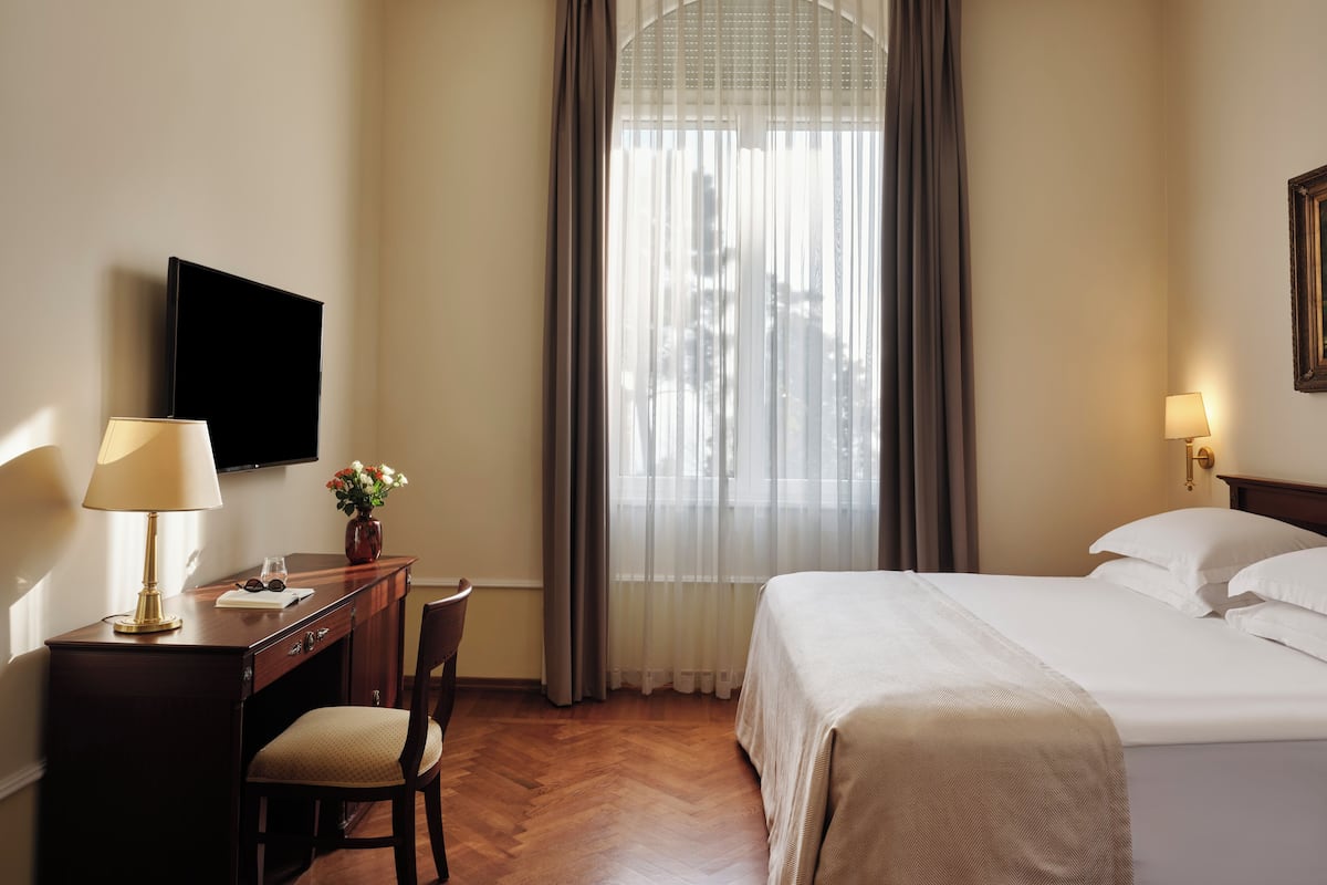 A hotel room with a bed, a desk with a lamp, a chair, a TV, and a window with curtains.