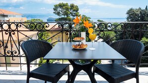 Un balcón con una mesa preparada para el desayuno, con cruasanes y un vaso de zumo de naranja, que ofrece vistas panorámicas del mar y edificios.
