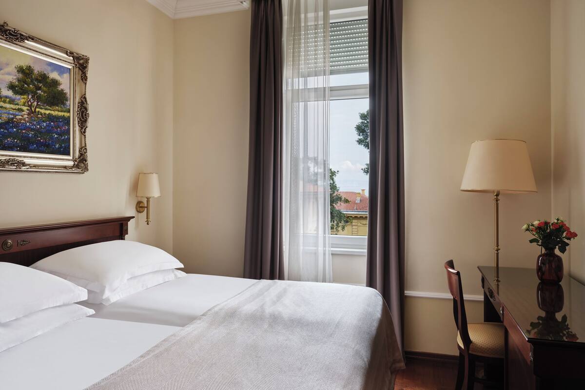 A hotel room with a large bed, a desk with a lamp, a chair, and a window with curtains.