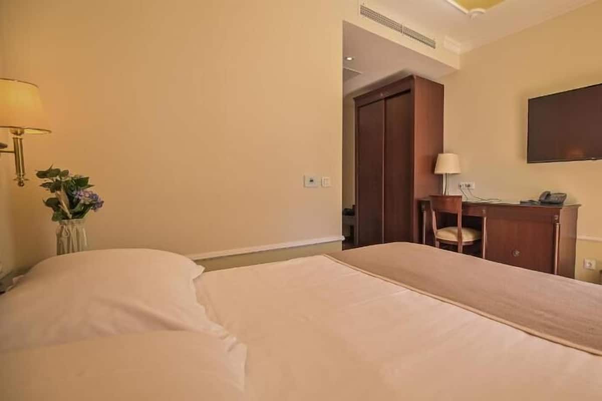 A hotel room with a bed, a desk with a lamp, a chair, a TV mounted on the wall, and a door leading to another room.