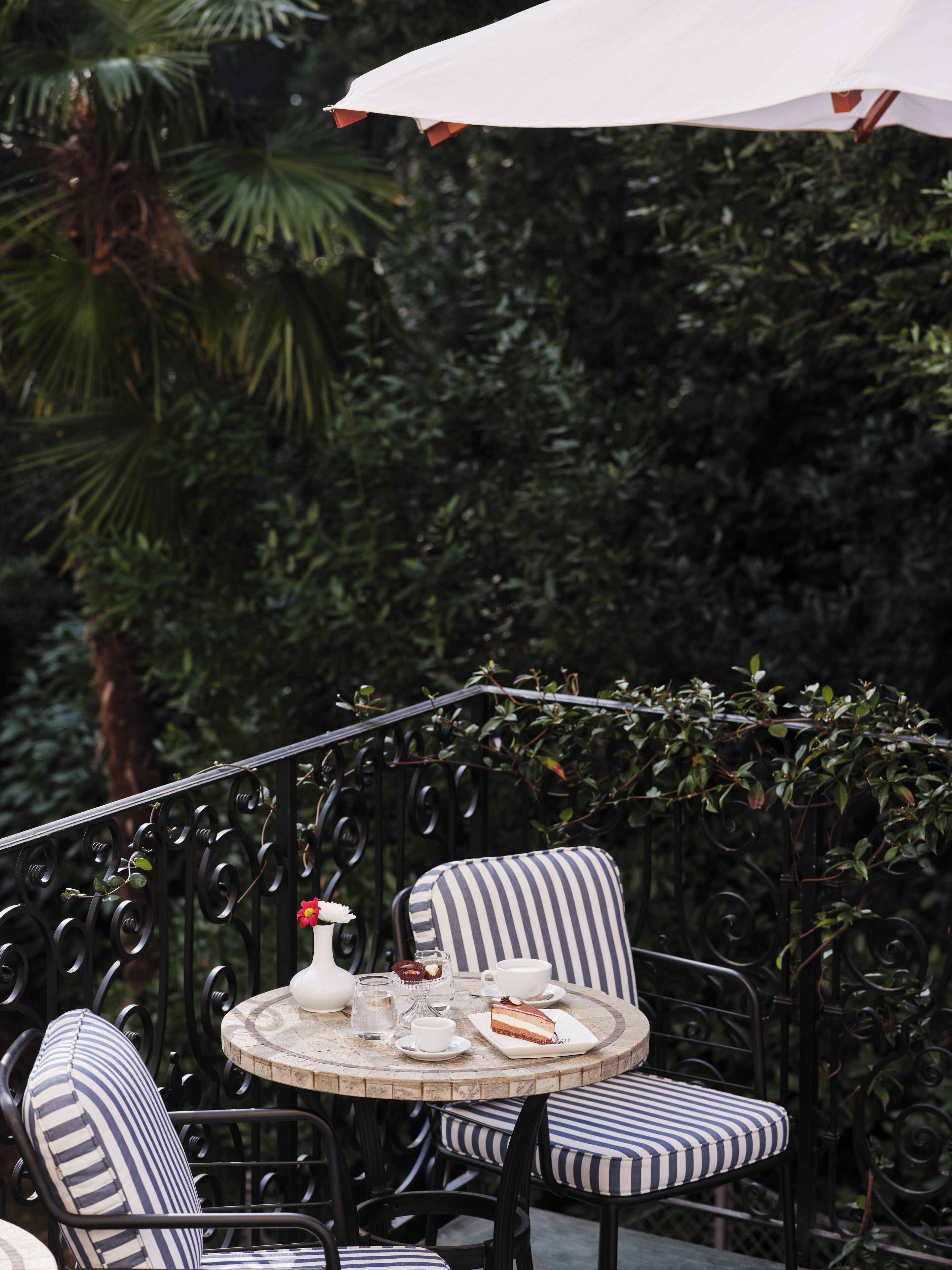 A round table with striped chairs, a white umbrella, and a small garden area with a metal fence and greenery.