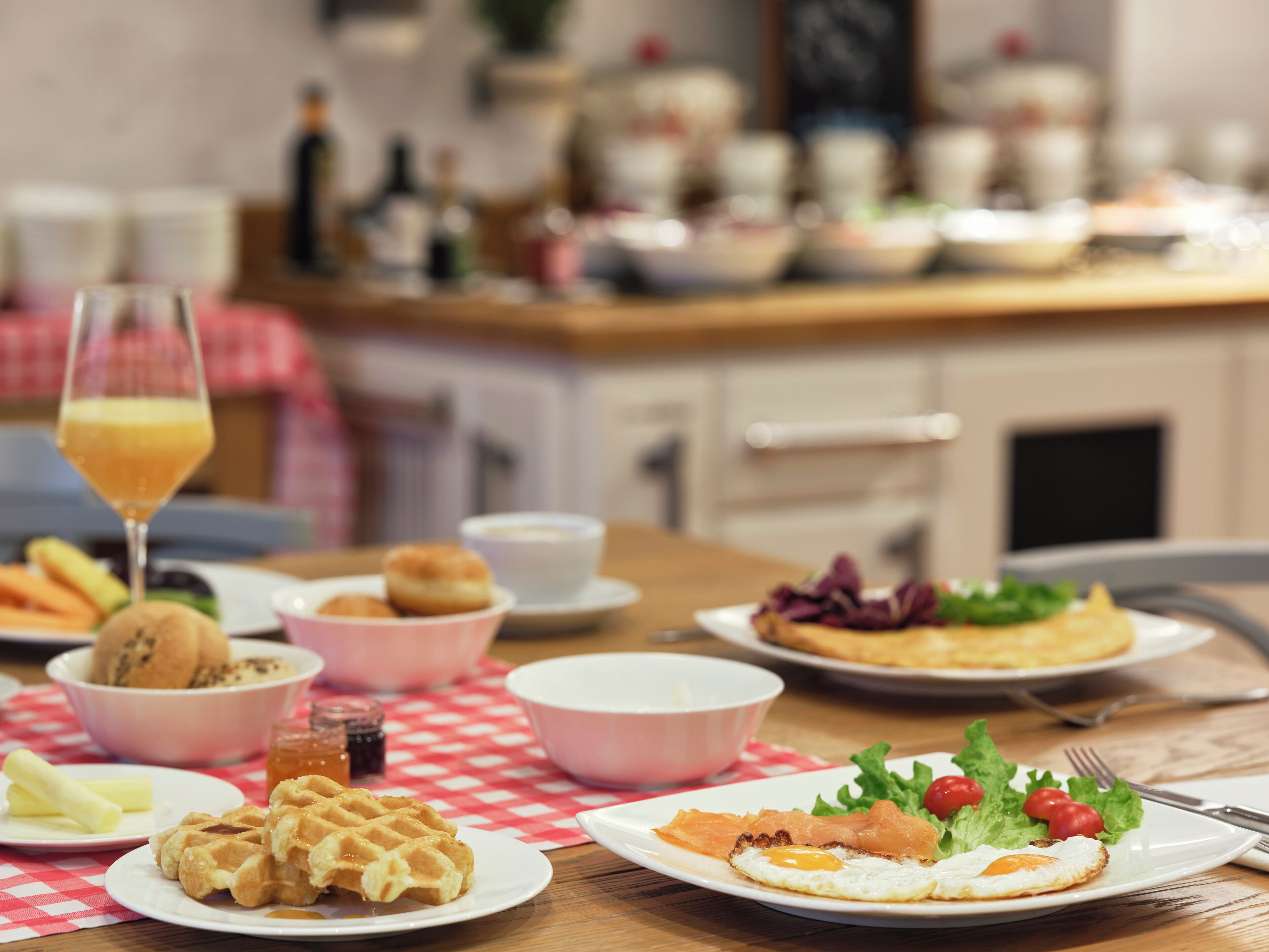 A breakfast setting with waffles, eggs, and salmon on plates, a glass of orange juice, and various bowls and cups on a checkered tablecloth.