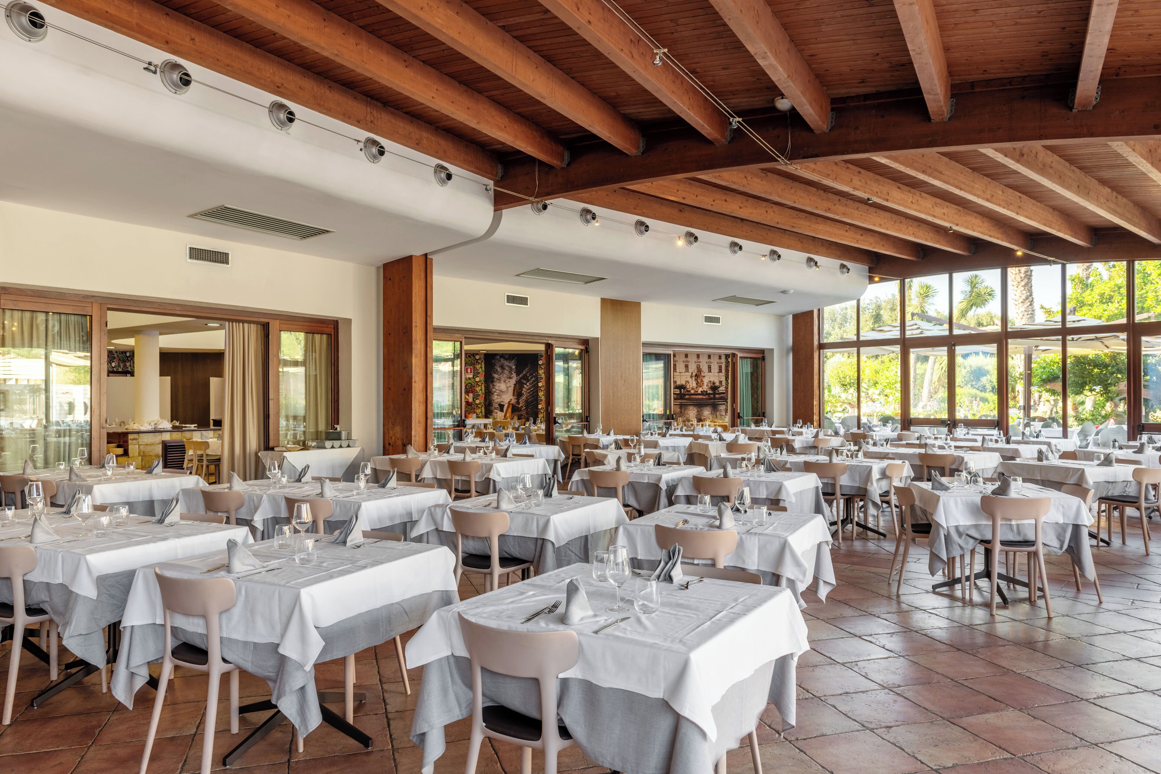 Un vaste espace dédié à la salle à manger, avec de nombreuses tables dressées, de grandes fenêtres laissant entrer la lumière naturelle et un plafond haut avec des poutres apparentes.