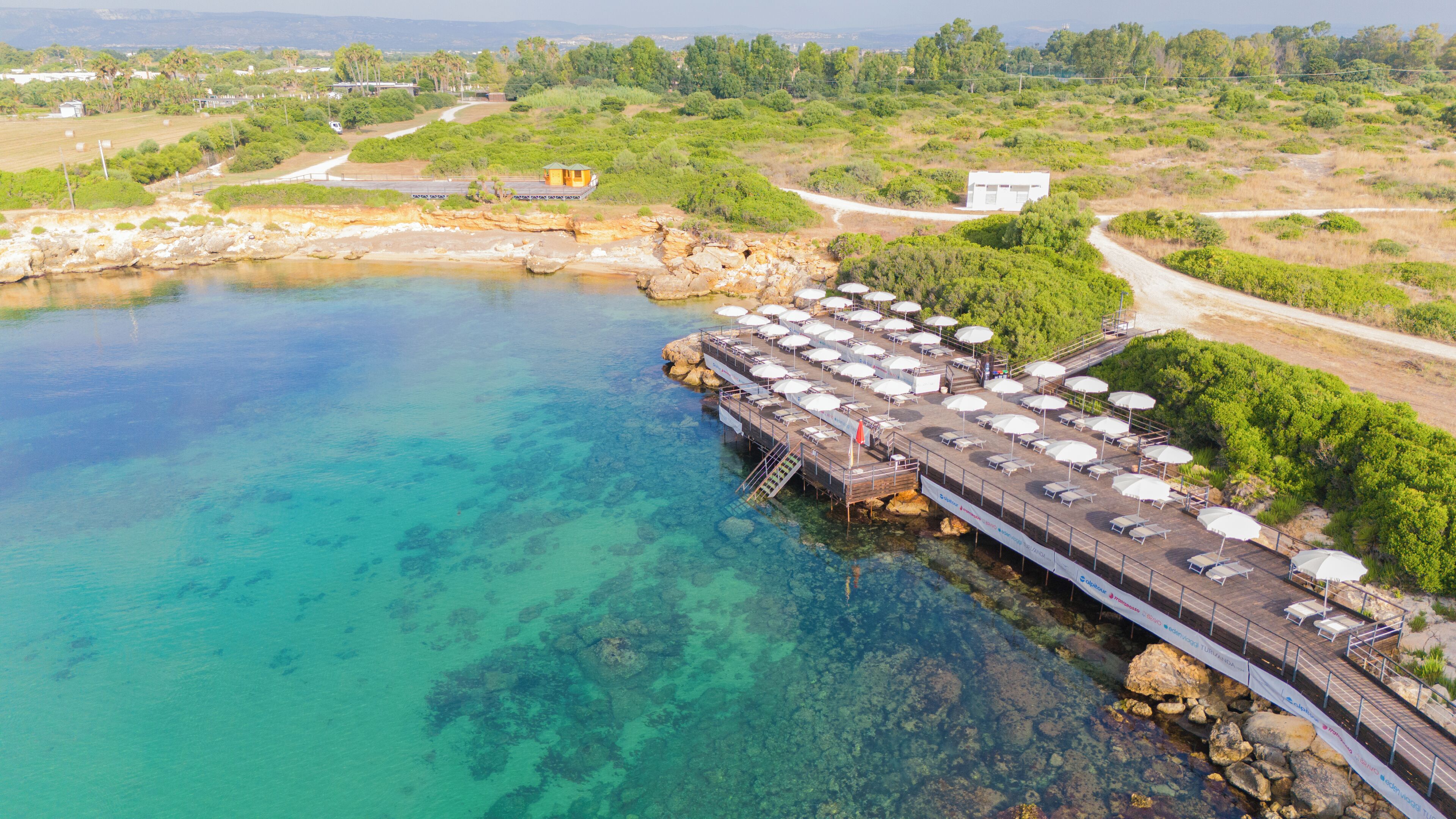 Luchtbeeld van een kustresort met een pier die zich uitstrekt over turquoise water, omringd door weelderige begroeiing en enkele gebouwen.