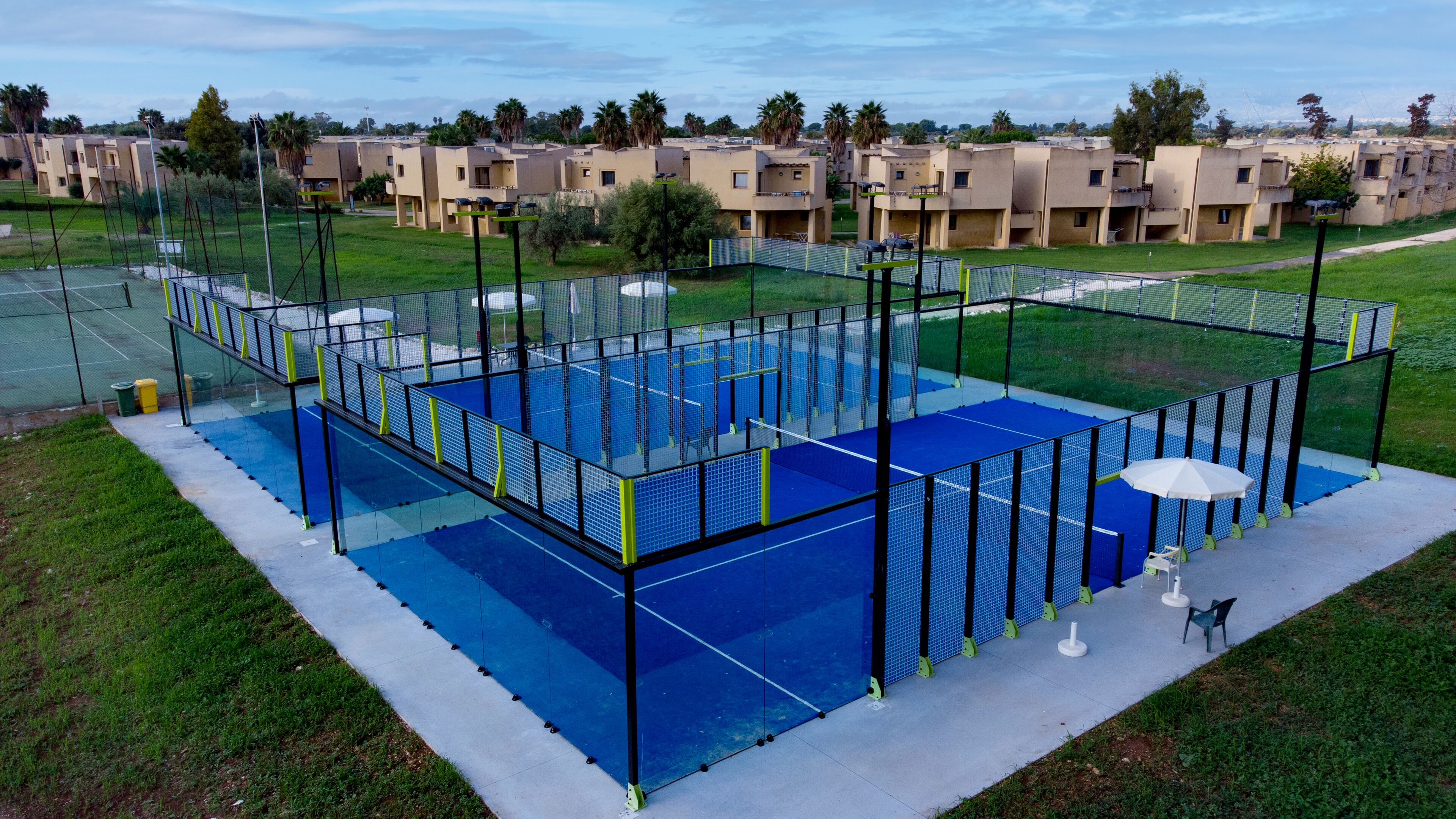 A tennis court surrounded by a fence, with a row of buildings in the background.