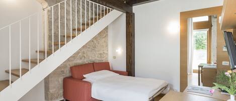 A bedroom with a stone wall, a wooden staircase, a red sofa, a bed, and a desk with a vase of flowers.
