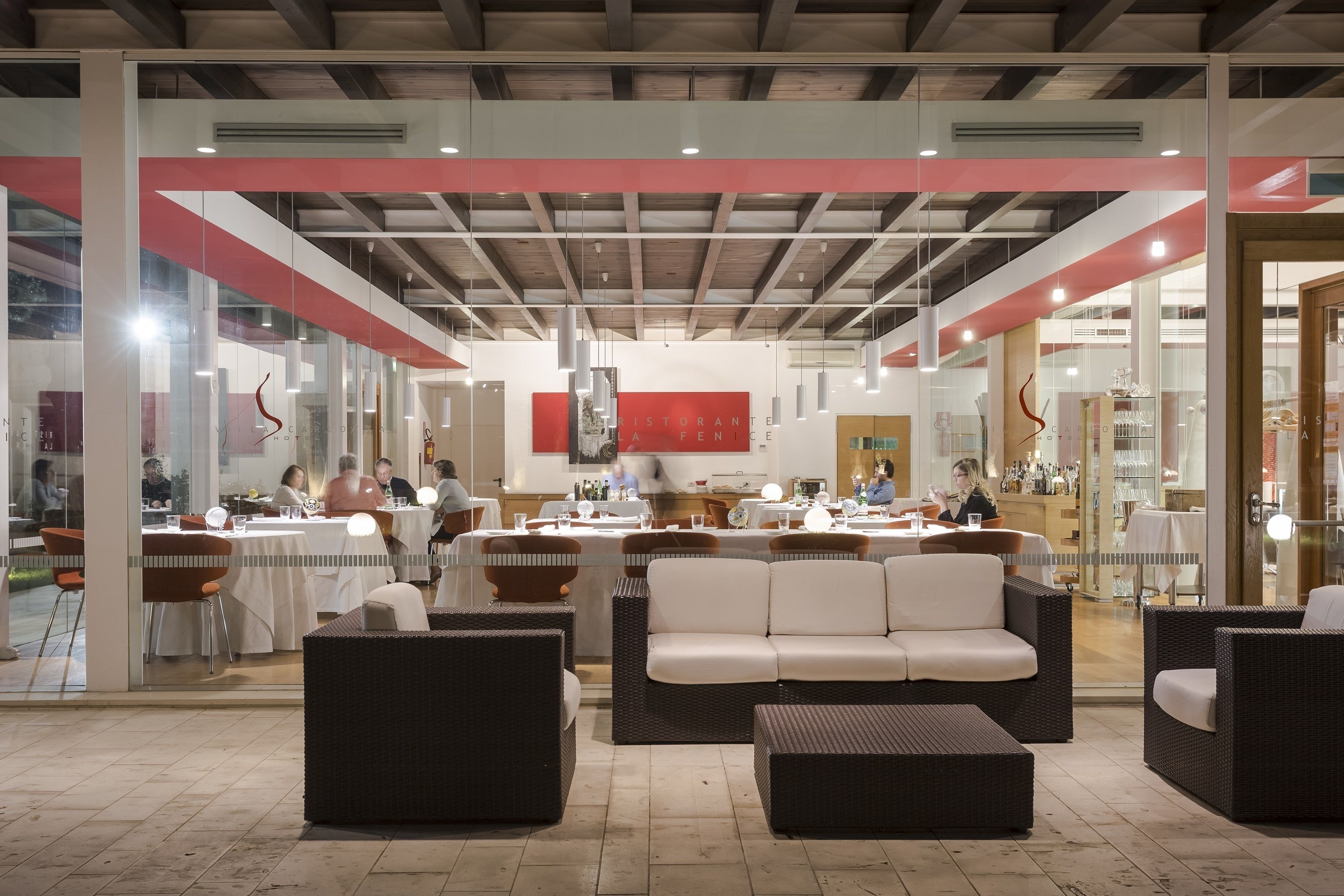 Un restaurant moderne doté d’un espace de salon central avec un canapé blanc et des fauteuils en osier, d’un bar avec des tabourets et d’une cuisine ouverte visible au fond.