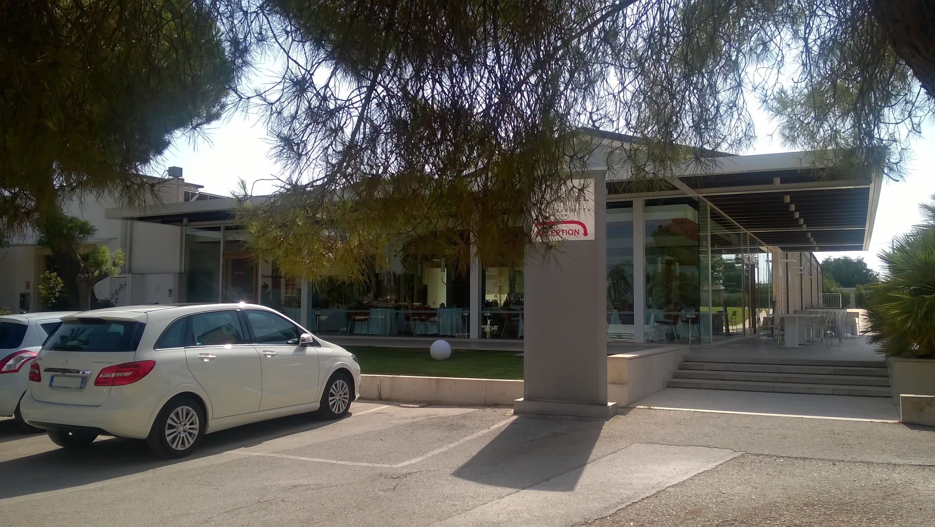 A modern building with a covered outdoor seating area, a white car parked in front, and a spherical object on the lawn.
