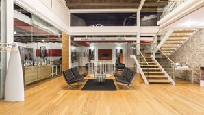 Amplio espacio tipo loft con techo alto, una mesa de vidrio central rodeada de sillas, una escalera con pared de piedra y una cocina moderna al fondo.