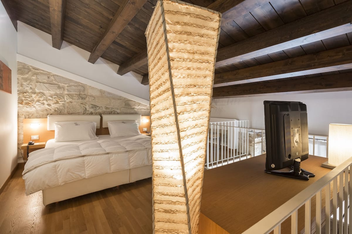 Une chambre avec un mur en pierre, un lit, une table de chevet, une lampe et un plafond en bois.