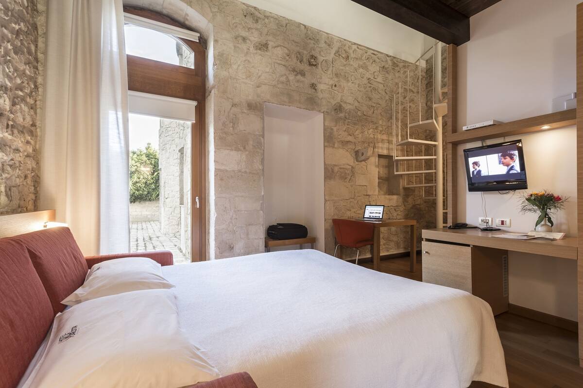 Une chambre d’hôtel avec un lit, un bureau, une télévision et un mur en pierre.