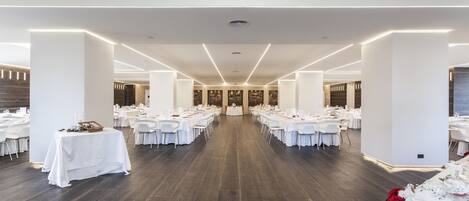 Un salón amplio con mesas y sillas blancas dispuestas en filas, suelo de madera oscura y paredes blancas.