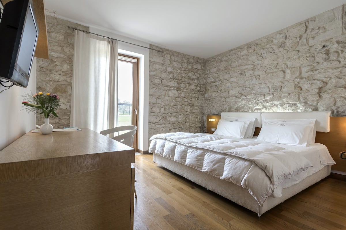 Une chambre avec un mur en pierre, un lit avec une literie blanche, un bureau avec une chaise et un téléviseur fixé au mur.