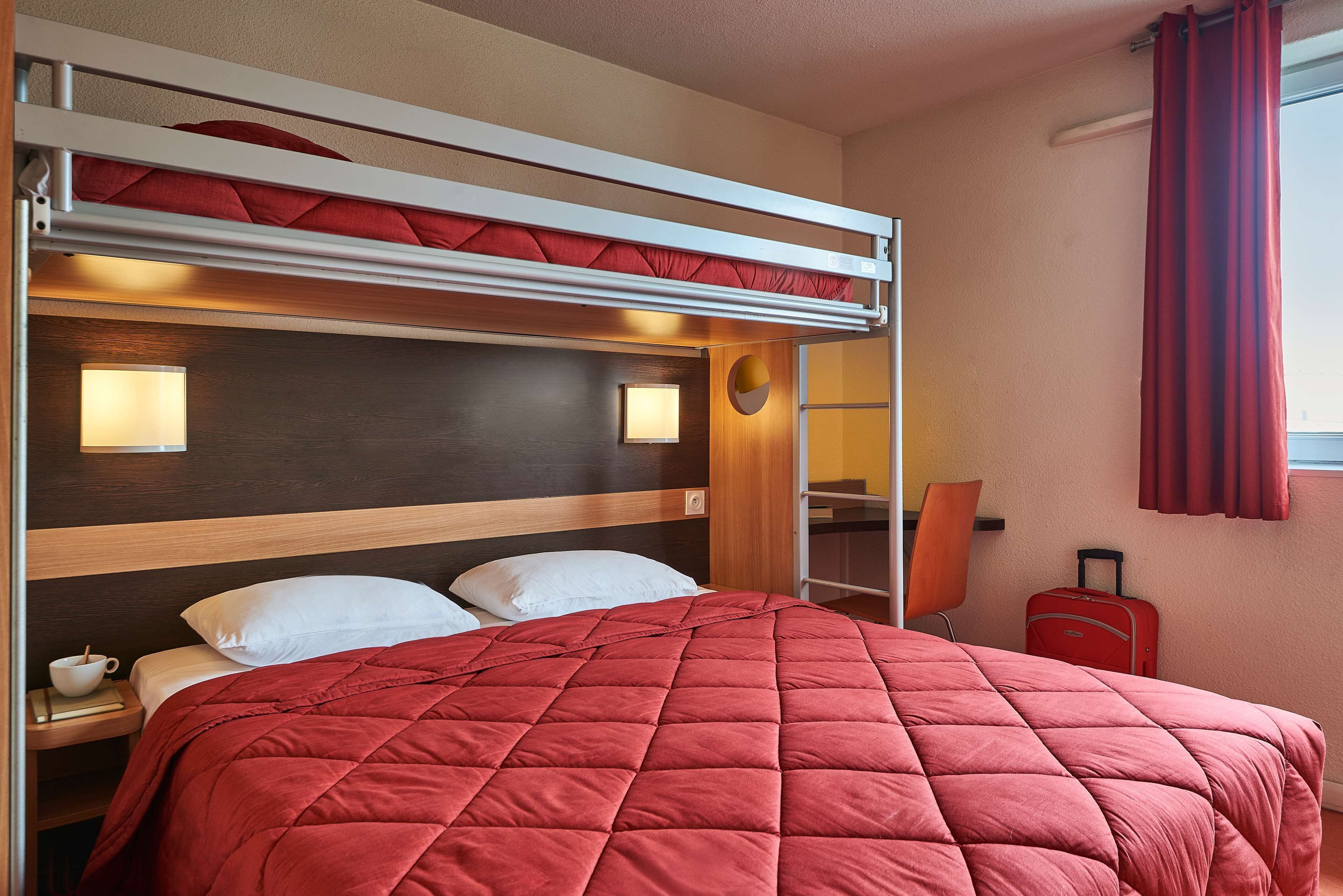 Un lit superposé avec une couverture matelassée rouge, des lampes de chevet et une valise dans une chambre d’hôtel.