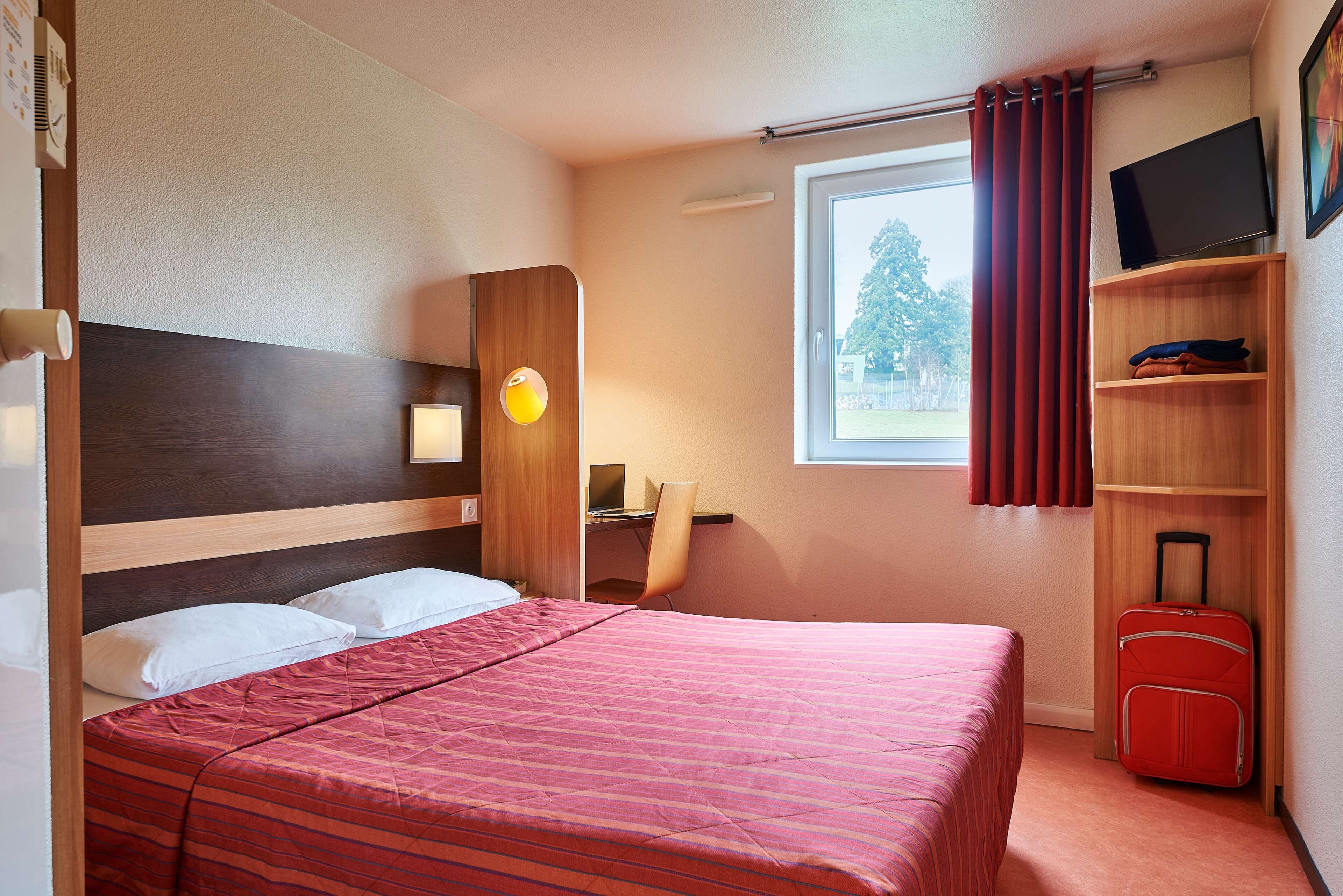 A hotel room with a bed, a desk, a chair, a TV, and a suitcase.