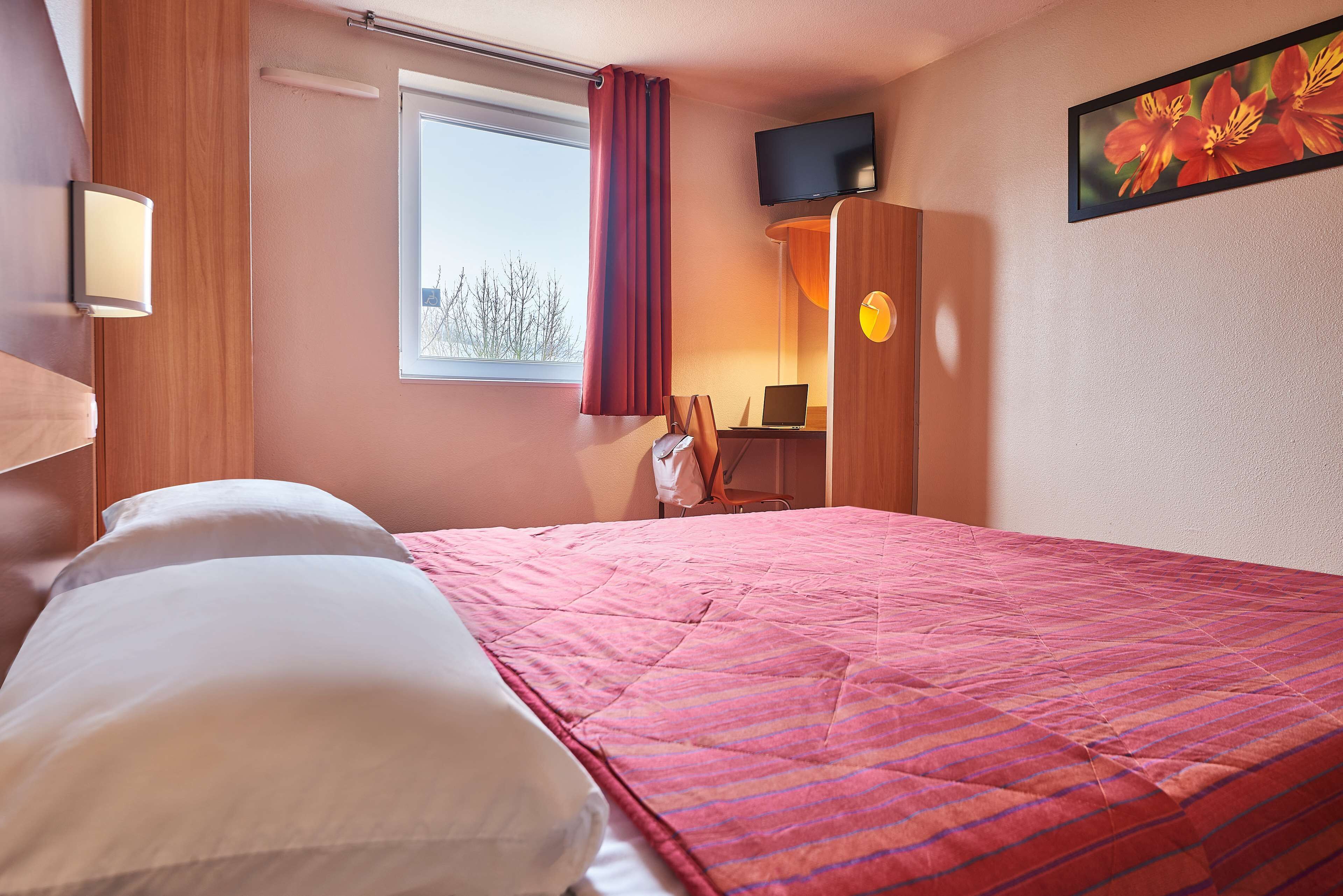 Una habitación de hotel con cama, escritorio, computadora portátil, televisión y ventana con cortinas.