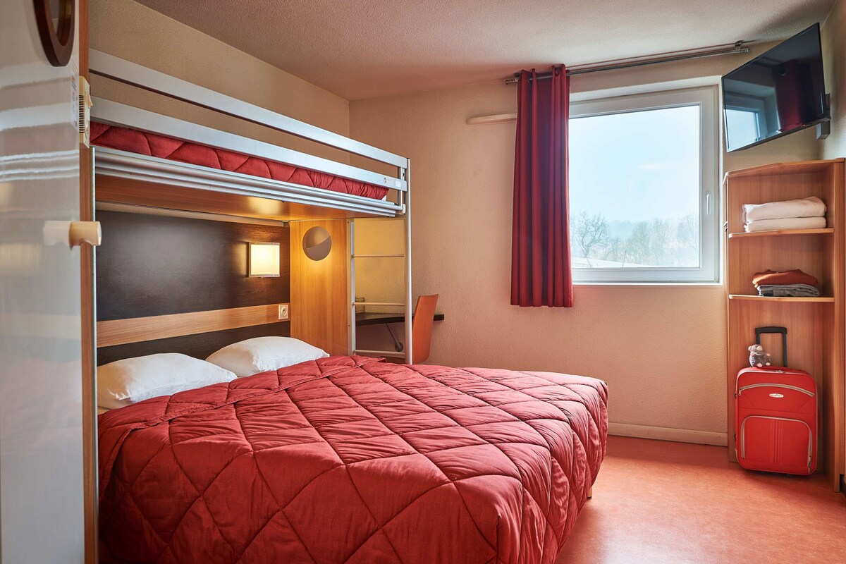 Una habitación pequeña con una litera, una cama individual, un escritorio, una ventana con cortinas, un estante con toallas dobladas y una maleta.