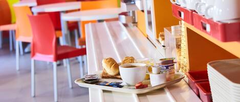 Bandeja de desayuno con croissants, café y mermelada sobre una mesa blanca en una cafetería de colores vivos.