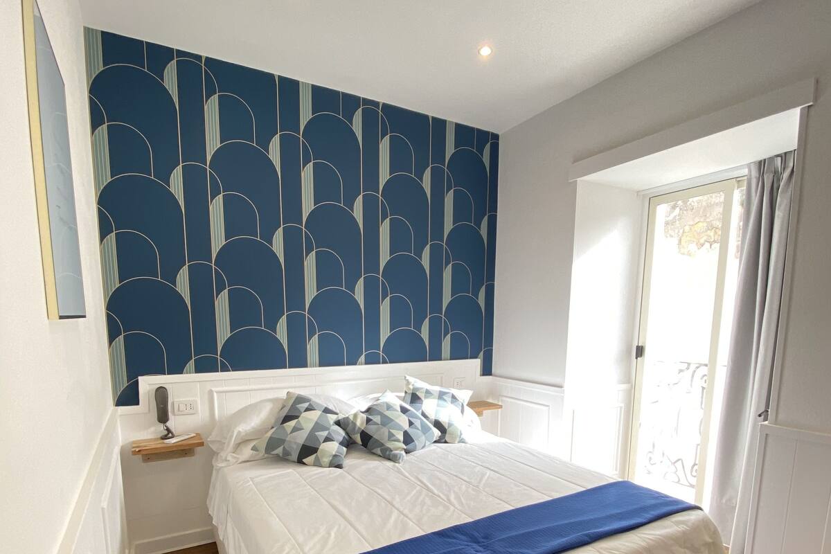 Une chambre à coucher avec un lit, une table de chevet, une commode, un mur à motifs bleus et une fenêtre avec des rideaux.