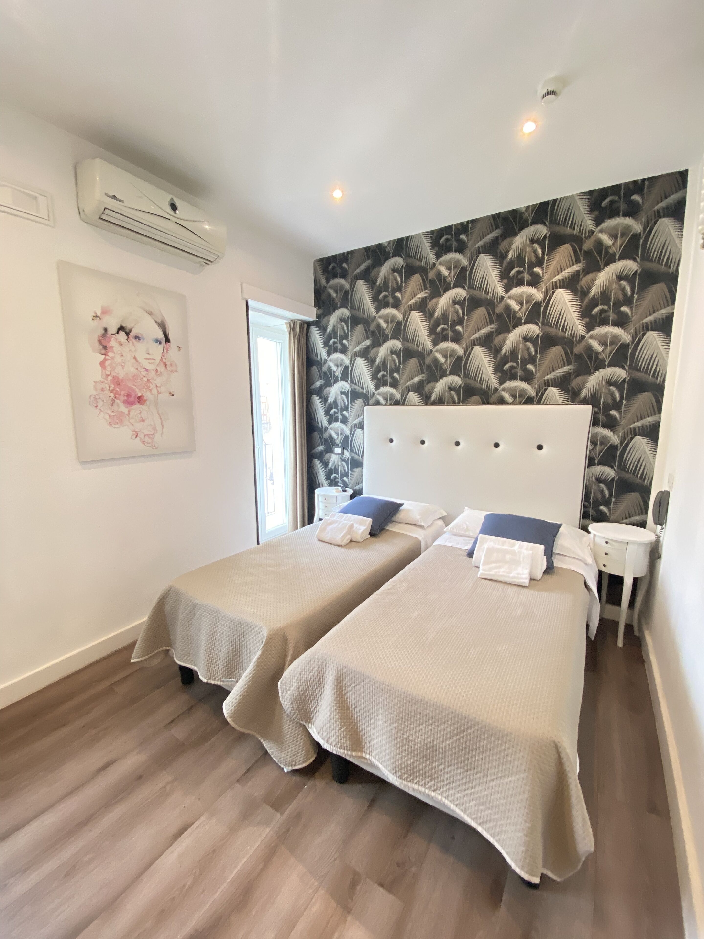Habitación de hotel con dos camas, un ventanal grande, un equipo de aire acondicionado, una obra de arte en la pared y un papel tapiz estampado.