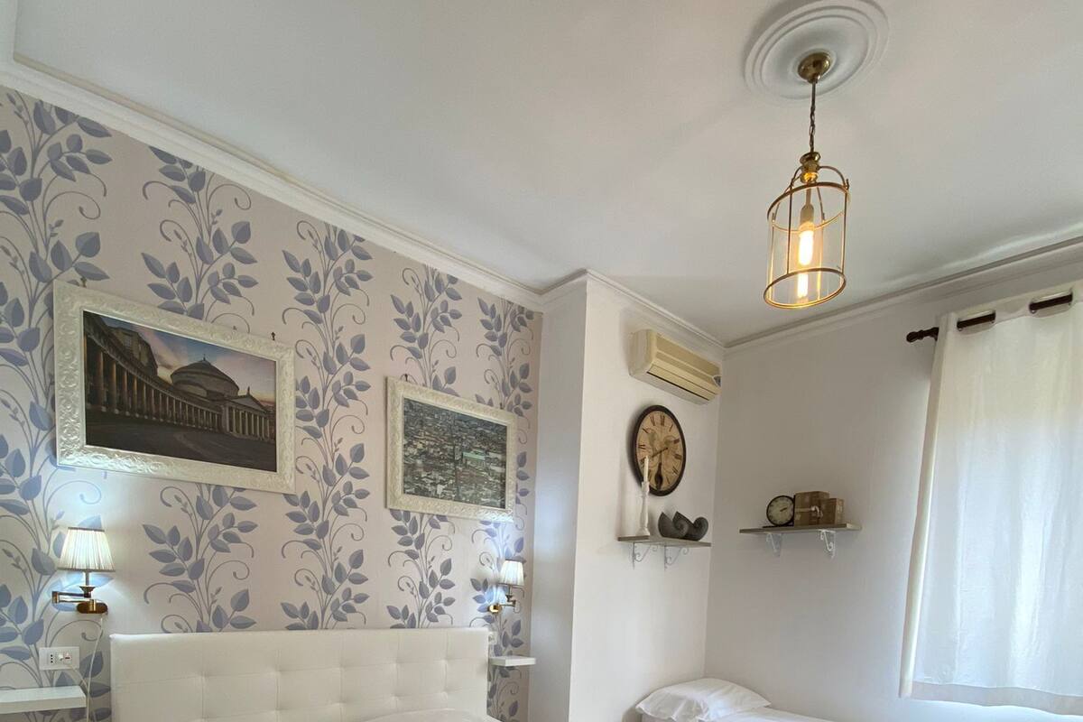 Une chambre à coucher avec un grand lit, un plus petit lit, une table de chevet, une lampe, une horloge et une étagère fixée au mur.