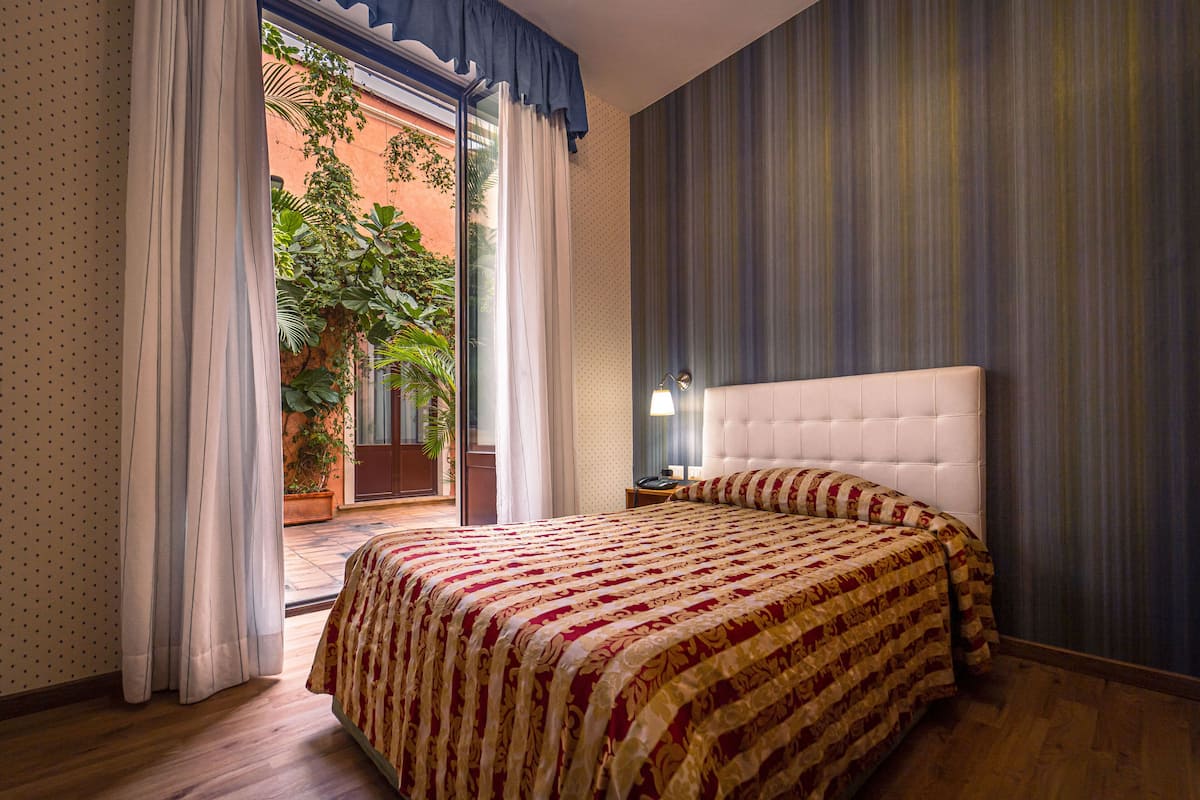 A bedroom with a large bed, a nightstand, a window with curtains, and a view of a garden.