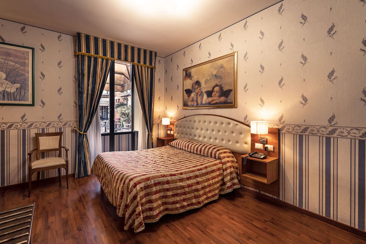 A hotel room with a large bed, two bedside tables with lamps, a chair, a window with curtains, and a painting on the wall.