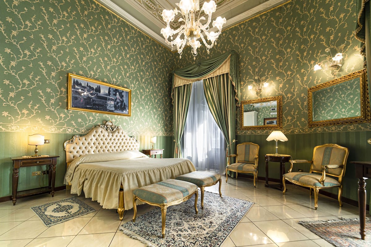 A luxurious bedroom with a large bed, two chairs, a bench, a chandelier, and a mirror.