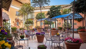 Un patio al aire libre con varias mesas y sillas, rodeado de macetas con plantas y sombrillas.