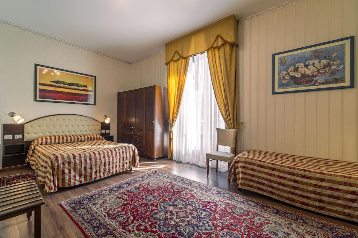 A hotel room with two beds, a large rug, a wooden wardrobe, and a painting on the wall.
