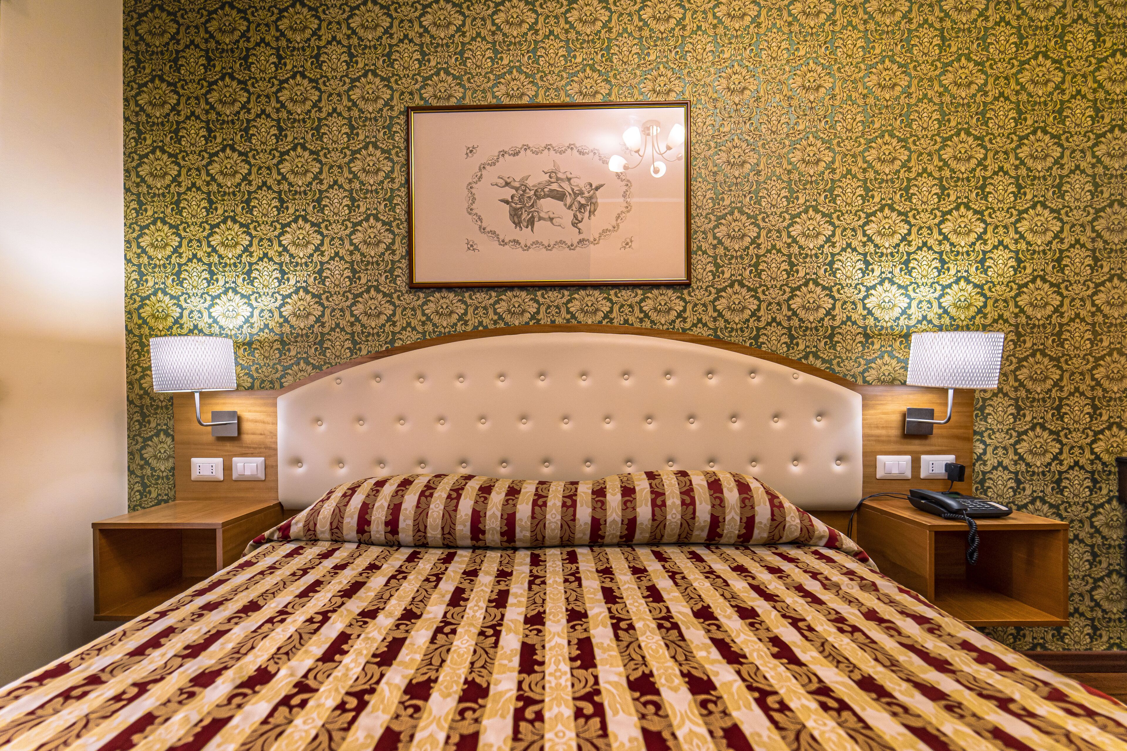 Een hotelkamer met een gestoffeerd hoofdeinde, een bed met een decoratieve sprei, twee bedlampen en een kunstwerk in een lijst aan de muur.