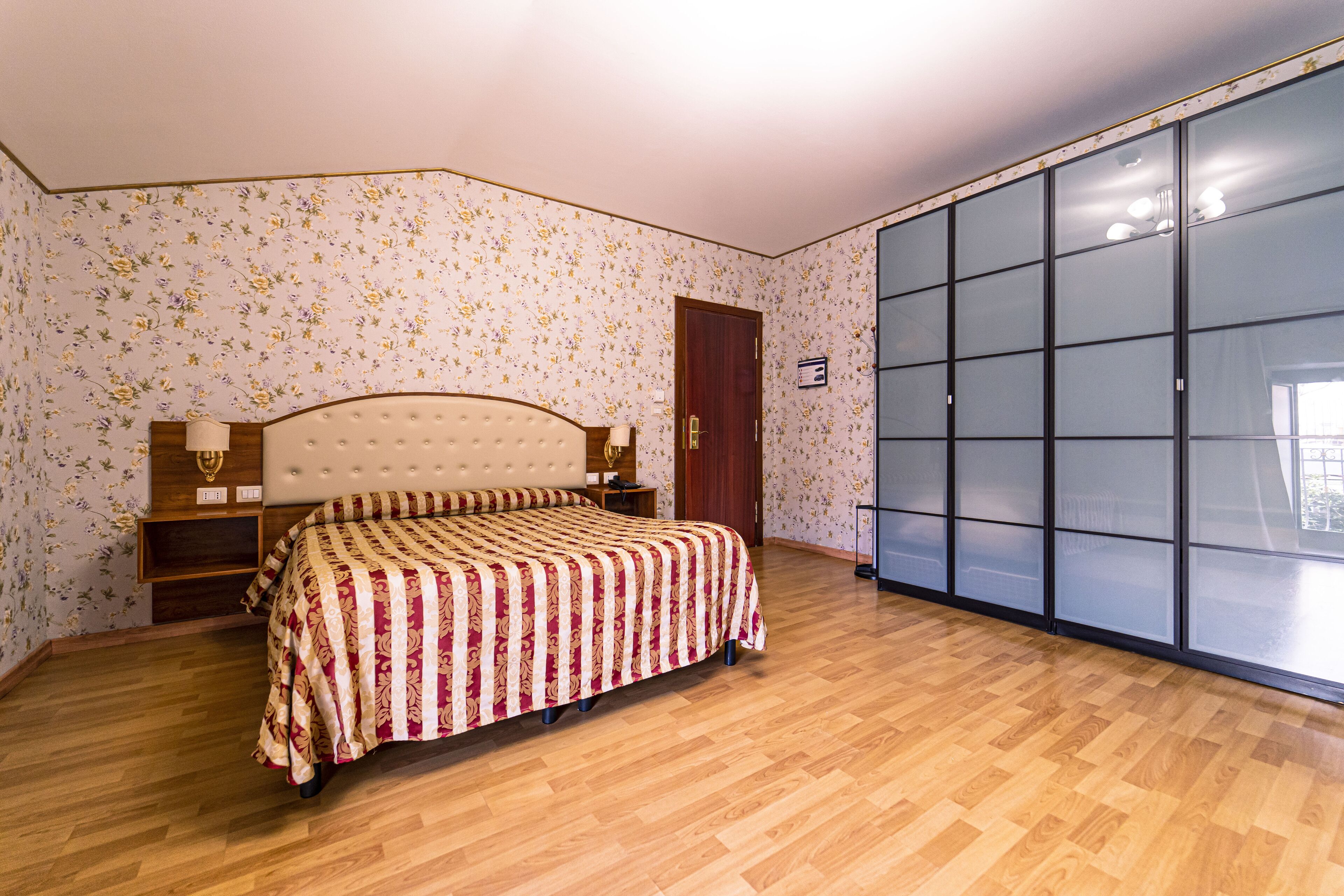 Une chambre à coucher avec un lit, un plancher en bois, un papier peint à motifs floraux, une armoire et une porte.