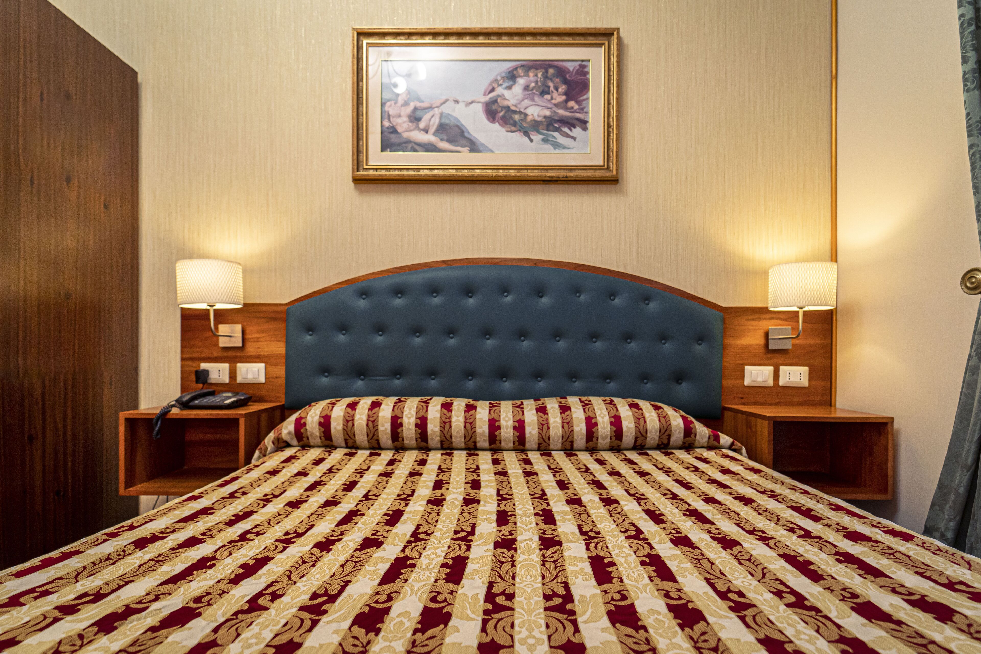 A hotel room with a large bed, two bedside tables with lamps, a wooden wardrobe, and a framed picture on the wall.