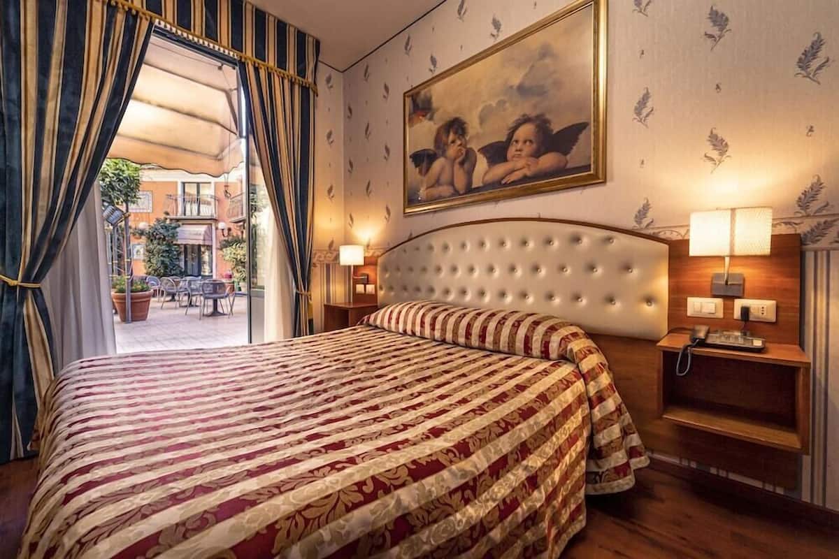 A hotel room with a large bed, a headboard, a painting, a lamp, and a view of the outside.