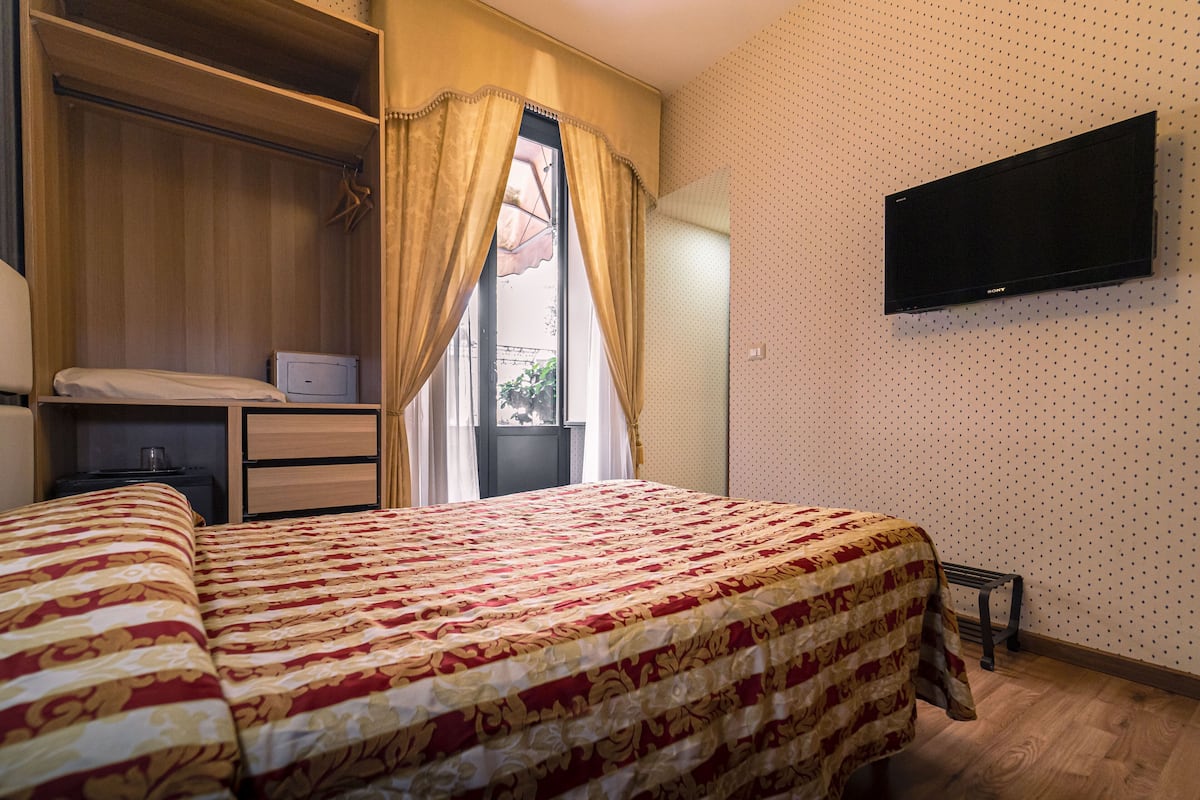 A bedroom with a bed, a wardrobe, a bedside table, a TV on the wall, and a window with curtains.
