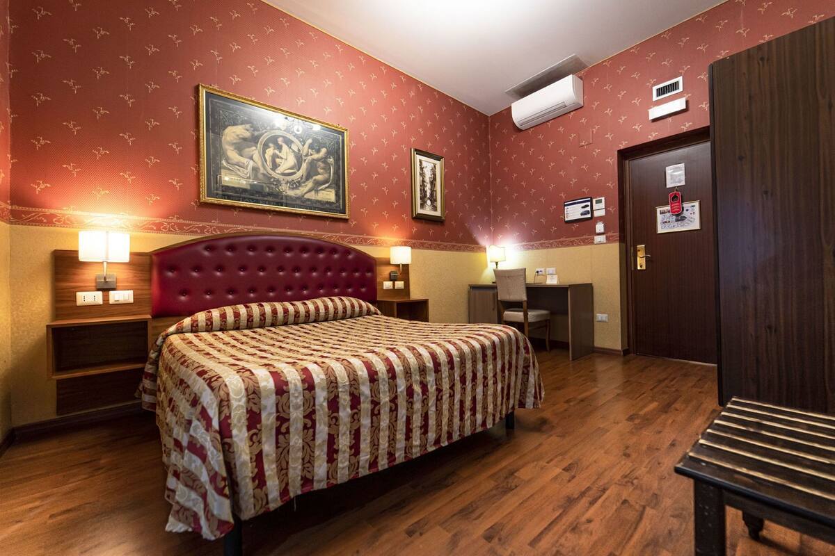 A hotel room with a large bed, a desk, a chair, a wardrobe, and two wall-mounted air conditioning units.