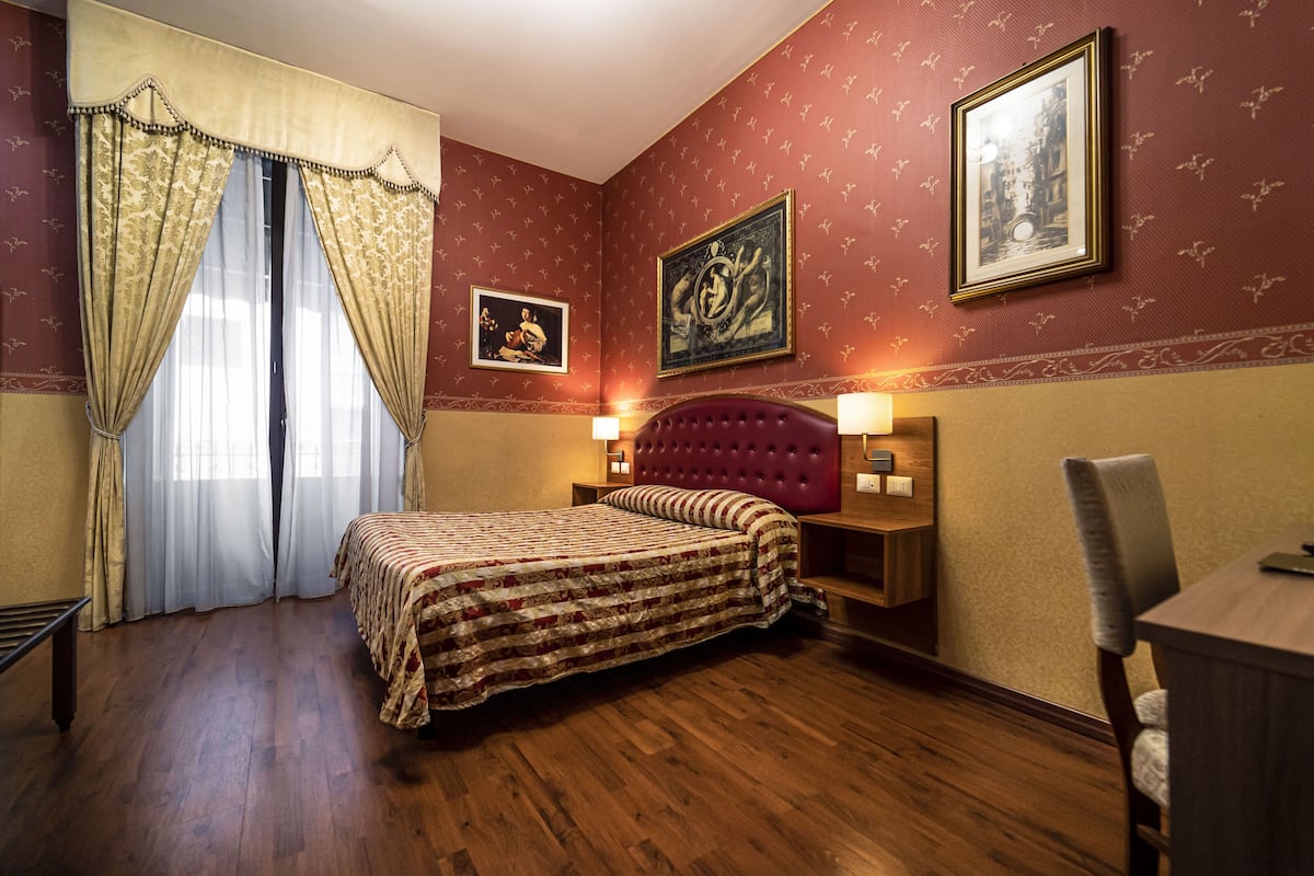 A hotel room with a large bed, a wooden floor, a red upholstered headboard, a nightstand with a lamp, and two framed pictures on the wall.