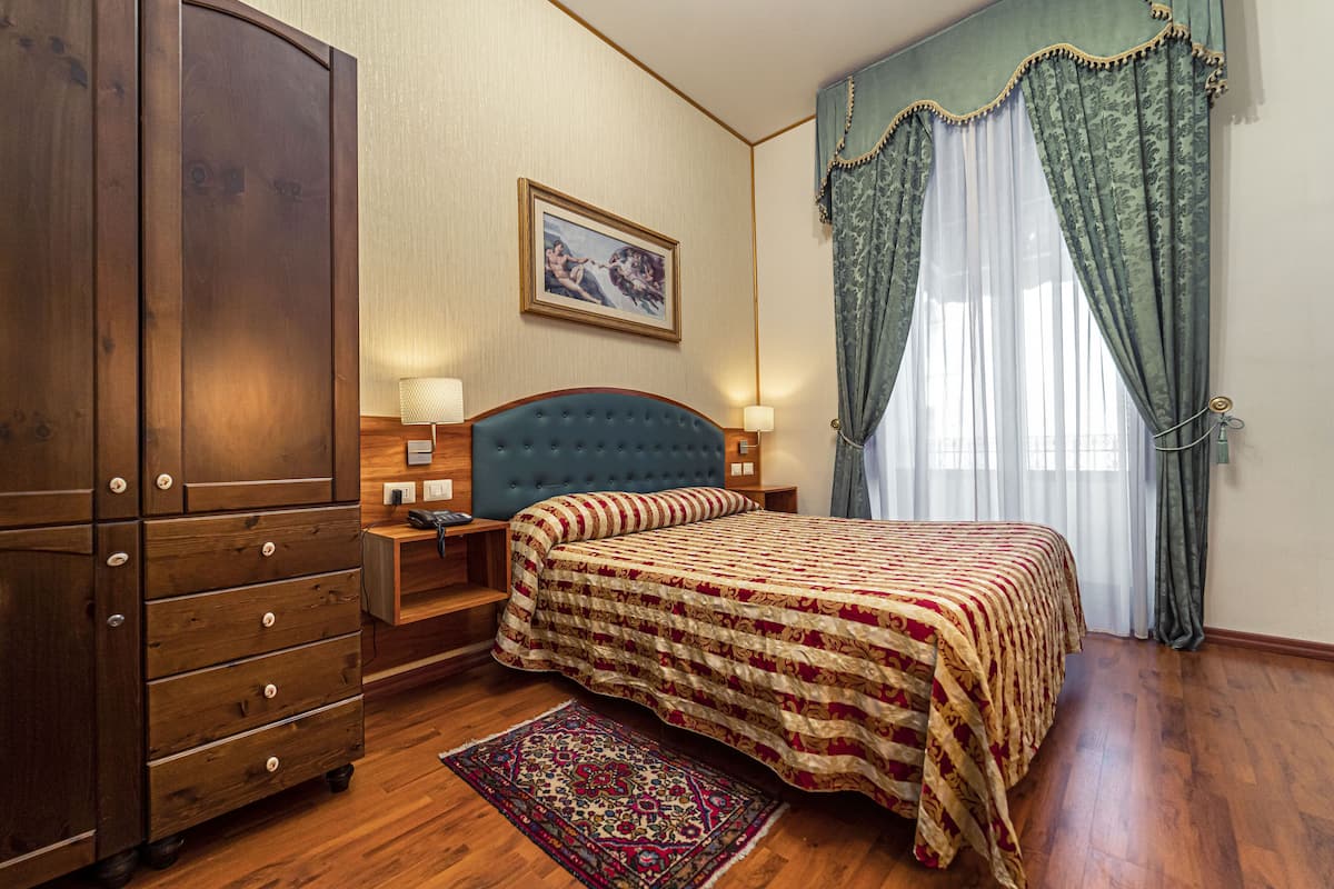 A bedroom with a large wooden wardrobe, a tufted headboard bed, a patterned bedspread, a nightstand with a lamp, and a window with curtains.